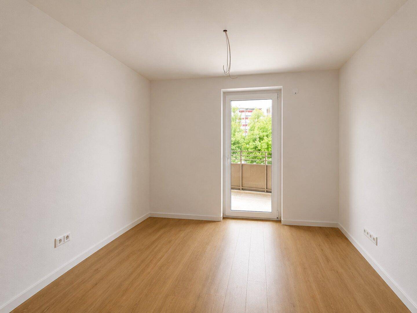 Helles, leeres Zimmer mit Laminatboden und Zugang zu einem kleinen Balkon.