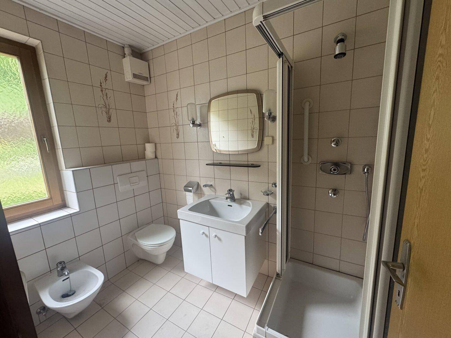 Gefliestes Badezimmer mit Dusche, WC, Bidet und Fenster, in gutem Zustand.