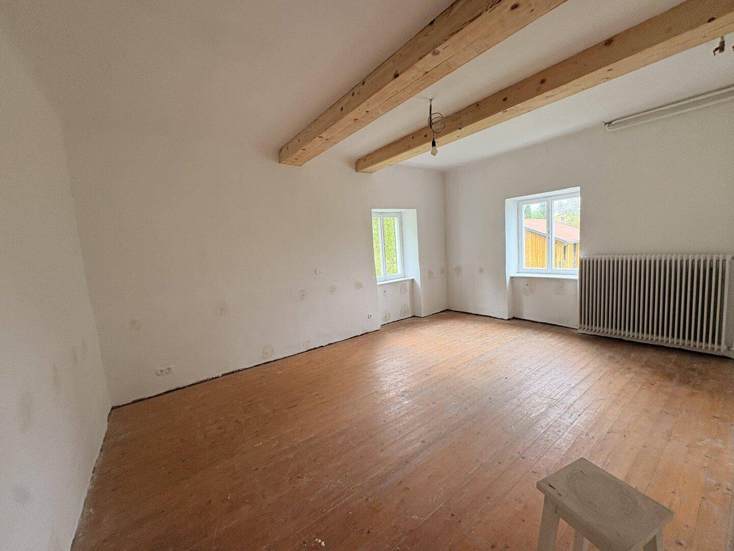 Heller Raum mit zwei Fenstern, Holzbalkendecke und Dielenboden, renovierungsbedürftig.