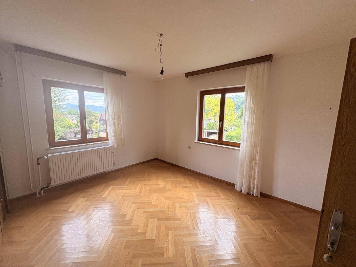 Schlafzimmer mit Parkettboden, zwei Fenstern und Blick ins Grüne.