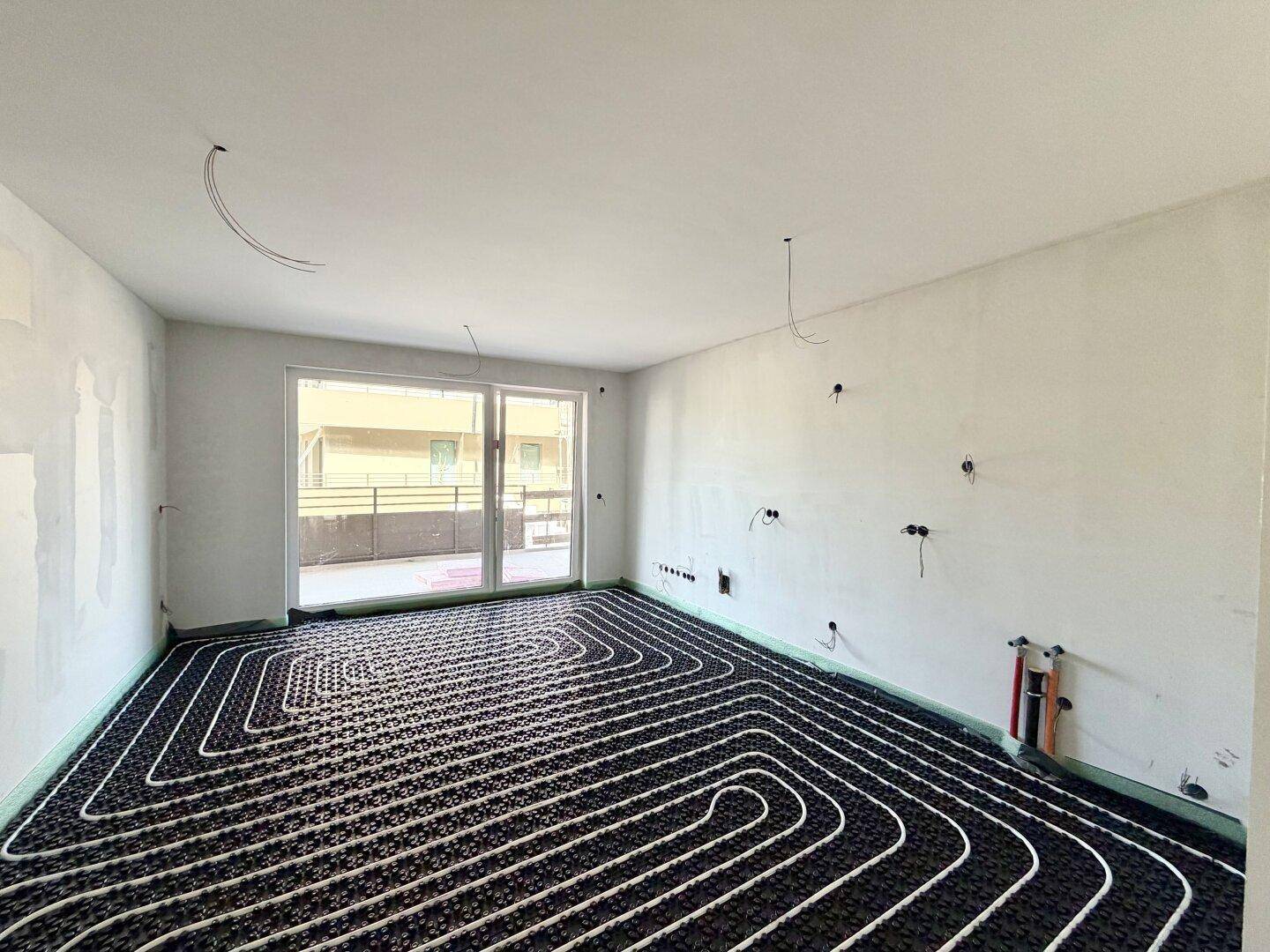Raum im Bau mit sichtbarer Fußbodenheizung und großen Fenstern, die zu einem Balkon führen.