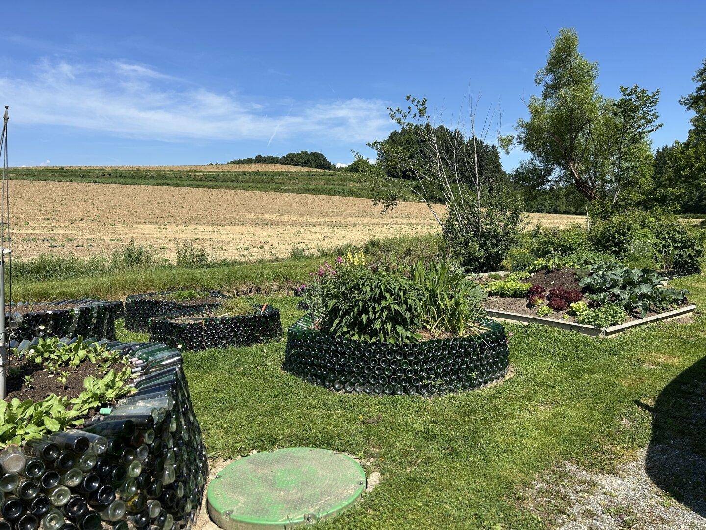 Kreativ gestalteter Gartenbereich mit Hochbeeten aus Glasflaschen und Blick auf Felder.