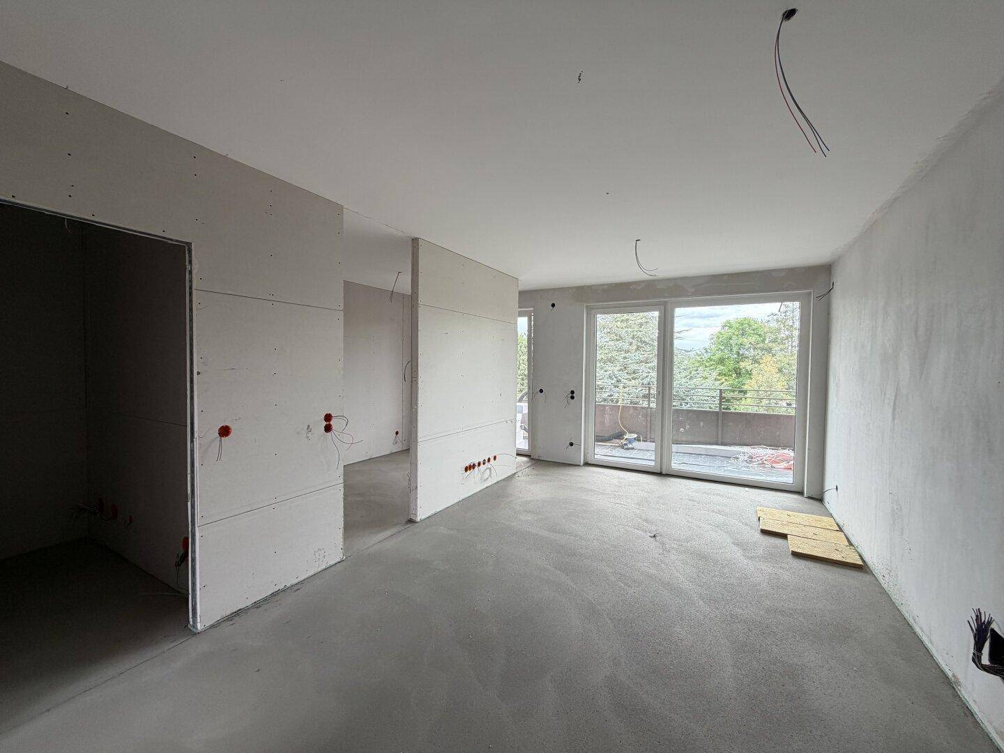 Rohbau eines Raumes mit Betonboden und großen Fenstern, die Zugang zu einem Balkon bieten.