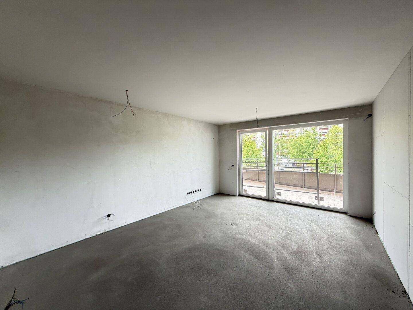 Unfertiger Wohnbereich mit Estrichboden und großen Fenstern, die auf einen Balkon führen.