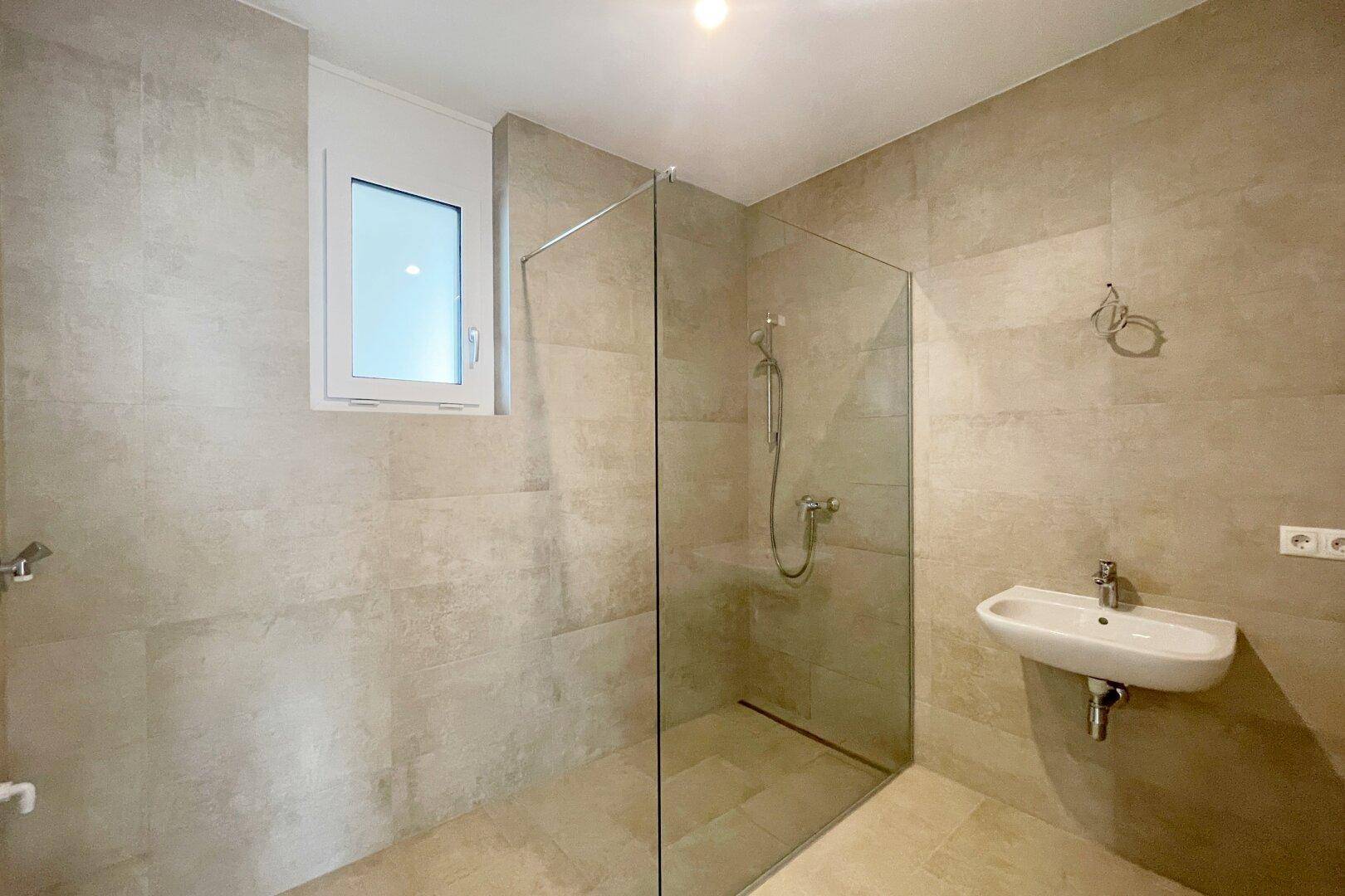 Modernes Badezimmer mit beigefarbenen Fliesen, ebenerdiger Dusche und Fenster für Tageslicht.