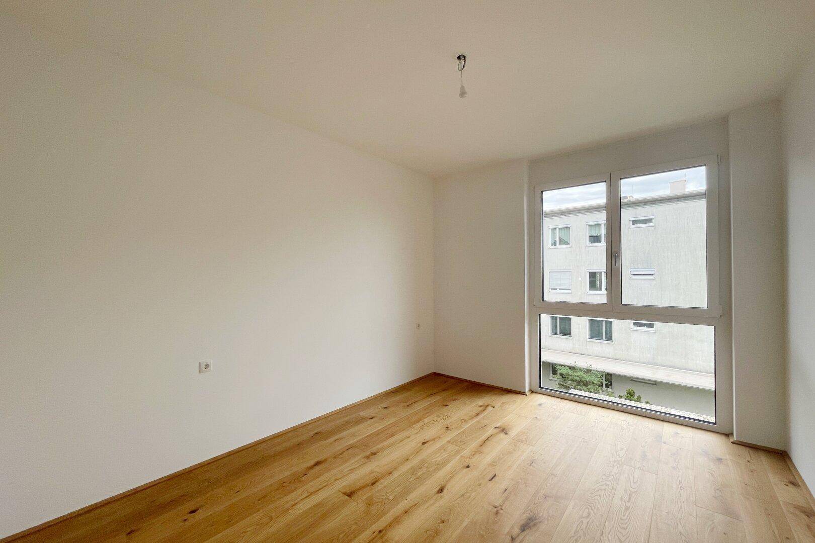 Freundliches Zimmer mit Holzfußboden und großem Fenster, ideal als Schlaf- oder Arbeitszimmer.