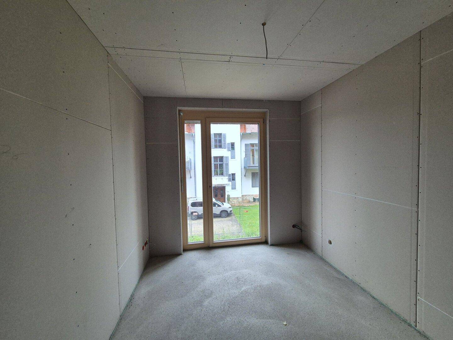 Raum im Rohbau mit Trockenbauwänden, Betonboden und großem Fenster mit Blick ins Grüne.