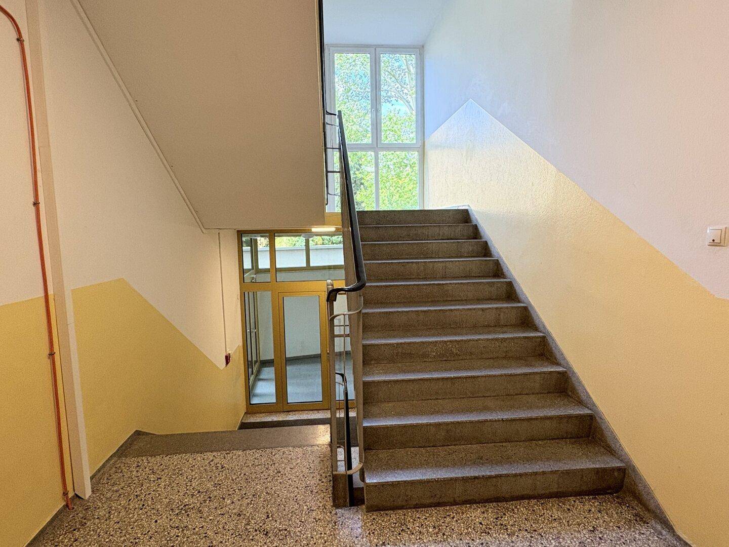 Helles Treppenhaus mit Steinstufen und großen Fenstern, die viel Tageslicht hereinlassen.