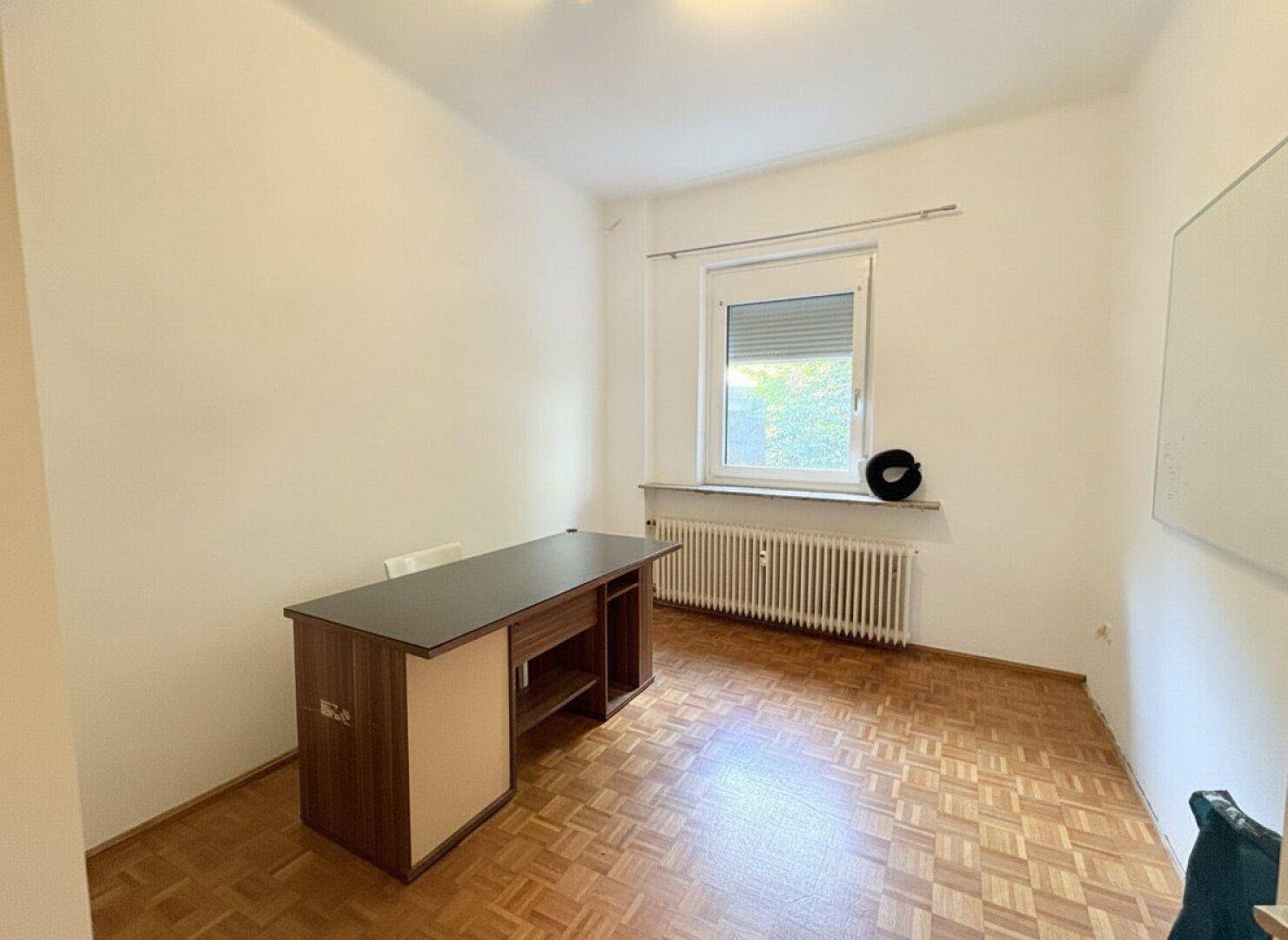 Helles Arbeitszimmer mit Parkettboden, Fenster und einem einfachen Schreibtisch.