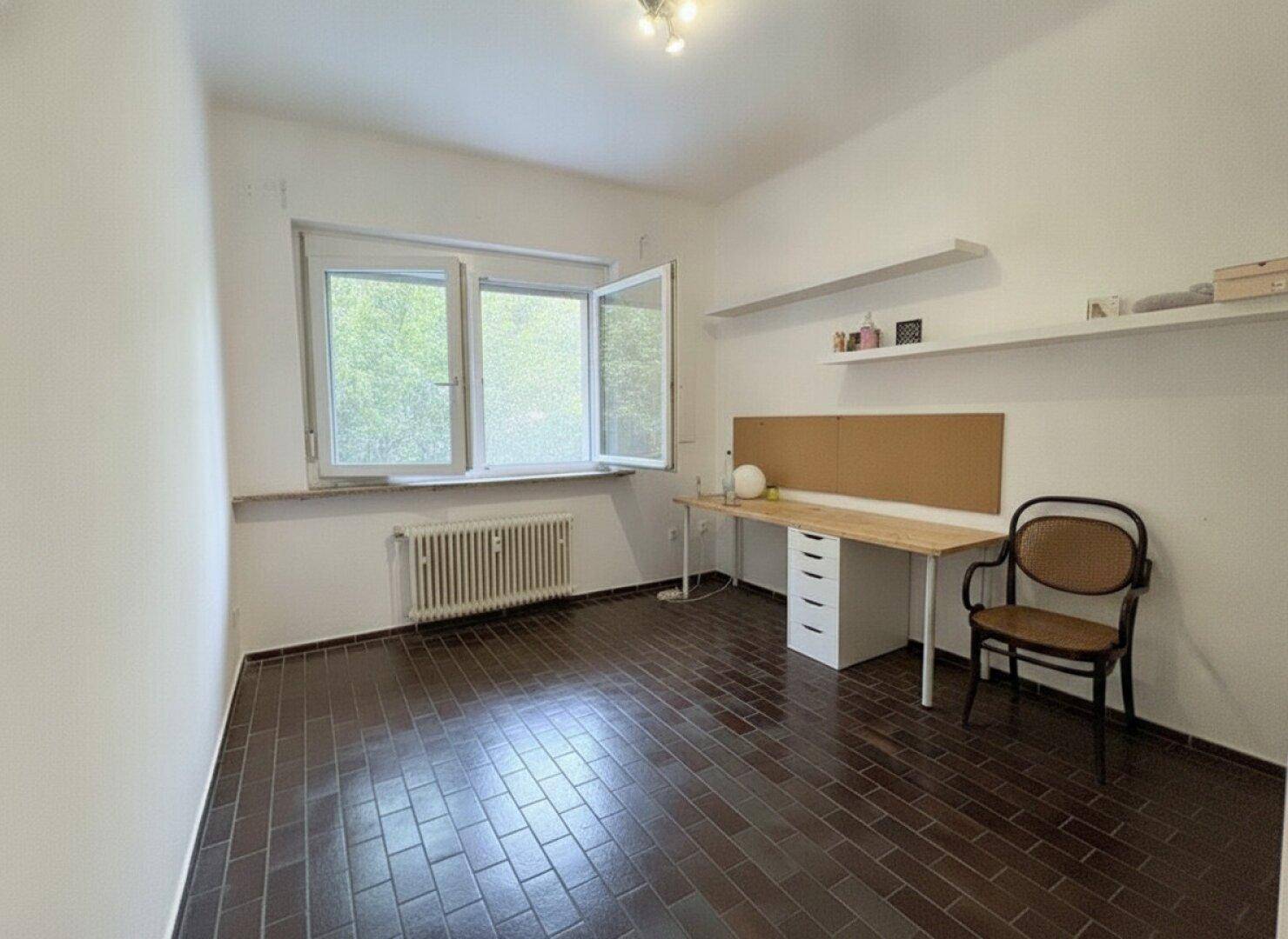 Geräumiges Zimmer mit dunklem Fliesenboden, Fenster und einem Arbeitsbereich mit Regalen.