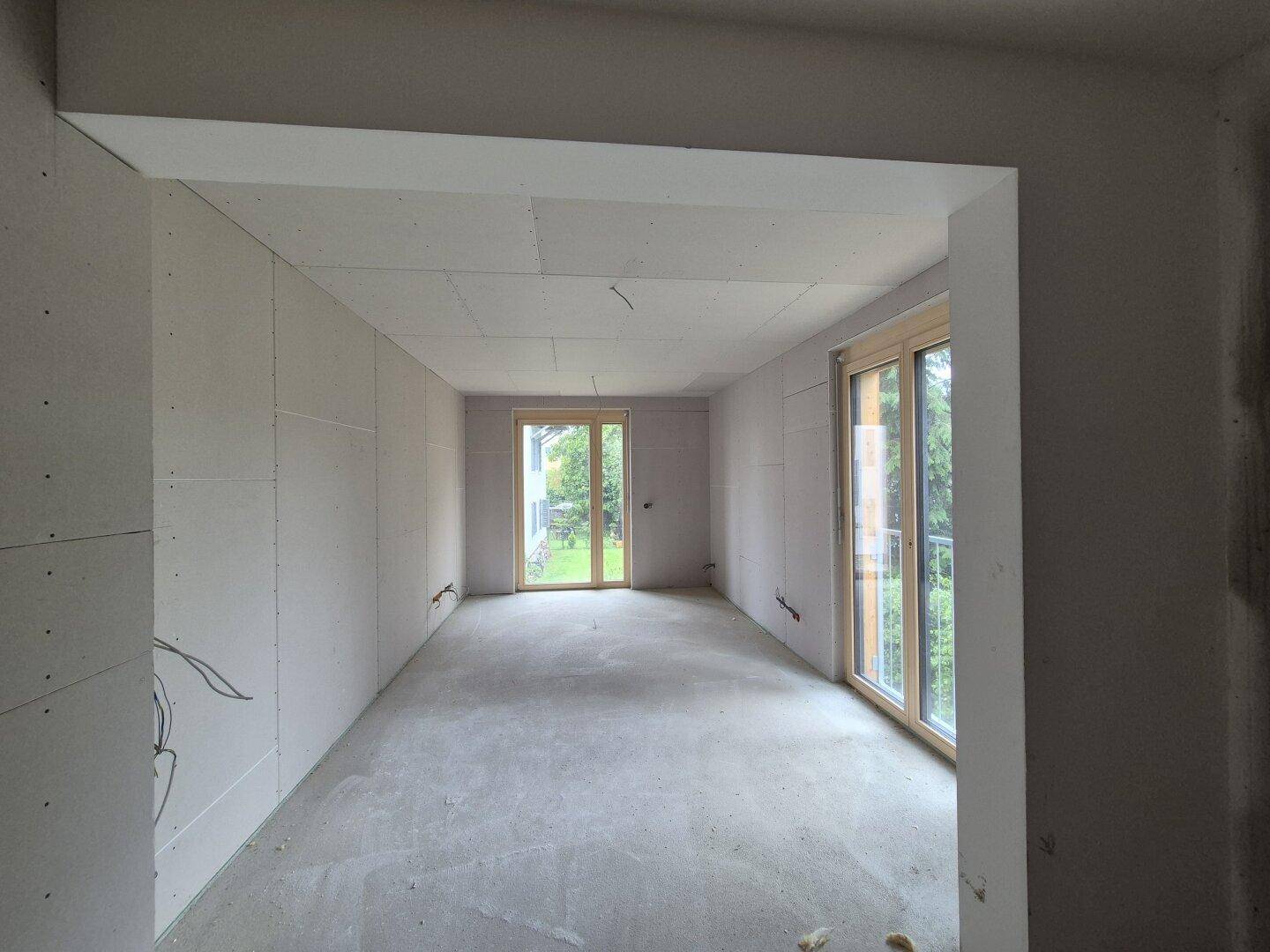 Rohbau eines Zimmers mit Gipskartonwänden und zwei großen Fenstern, bereit für den Innenausbau.