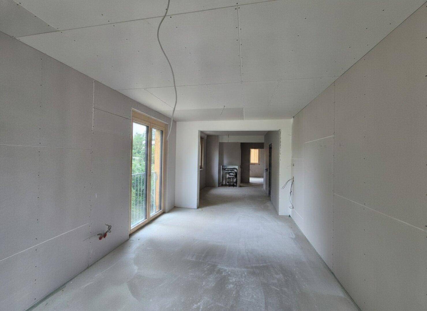 Zimmer im Rohbau mit Gipskartonwänden und großen Fenstern, die viel Tageslicht hereinlassen.