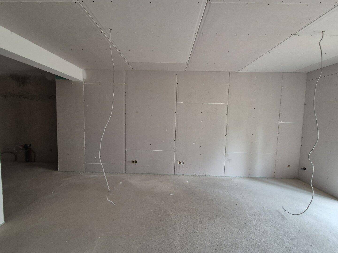 Leerer Raum im Rohbau mit Gipskartonwänden und hängenden Kabeln, wartet auf Fertigstellung.
