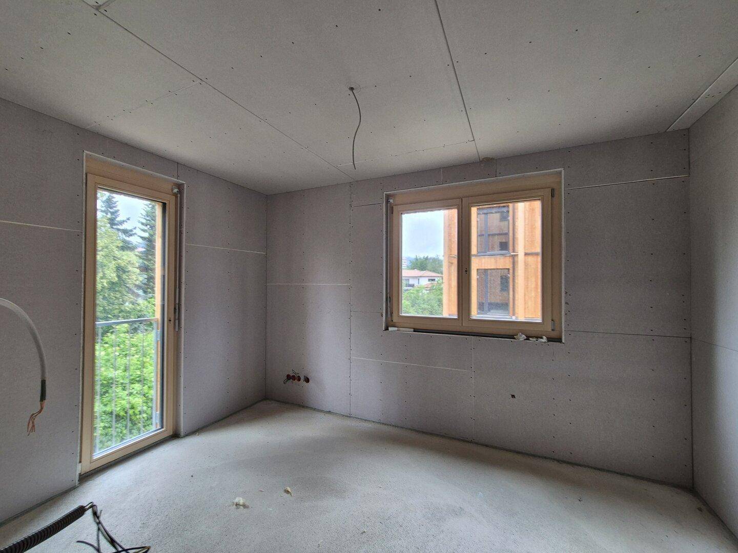 Rohbau eines Zimmers mit Gipskartonwänden und zwei Fenstern, die einen Blick ins Grüne bieten.