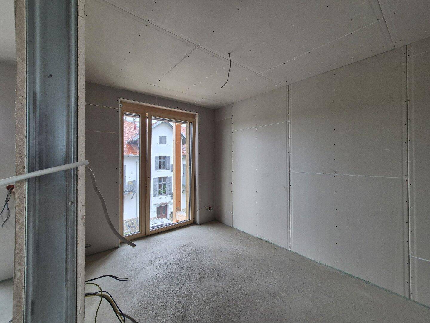 Rohbau eines Zimmers mit Betonboden und Fenster, das auf die umliegenden Gebäude blickt.