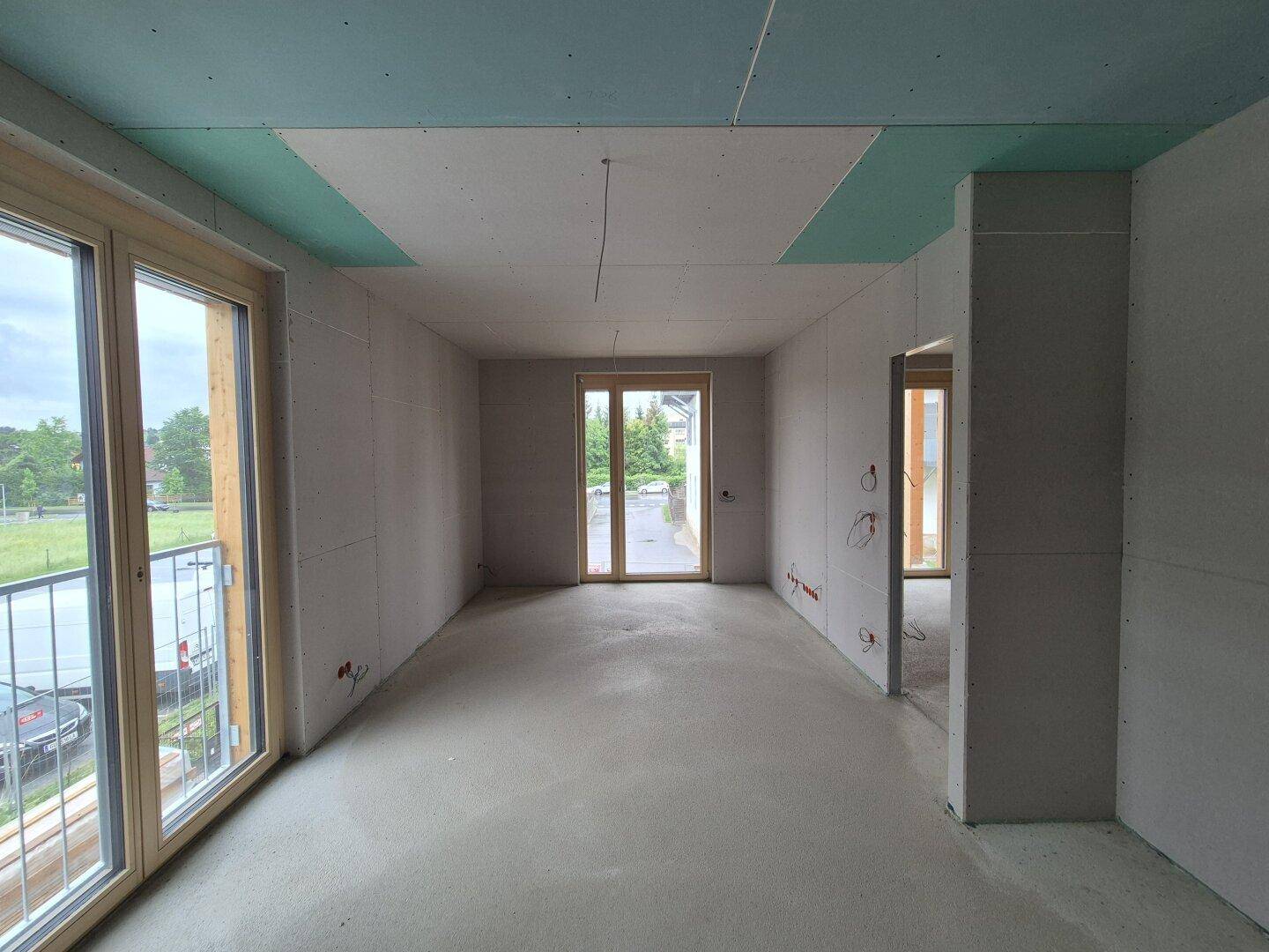Rohbau eines Wohnzimmers mit Betonboden, Trockenbauwänden und großen Fenstern.