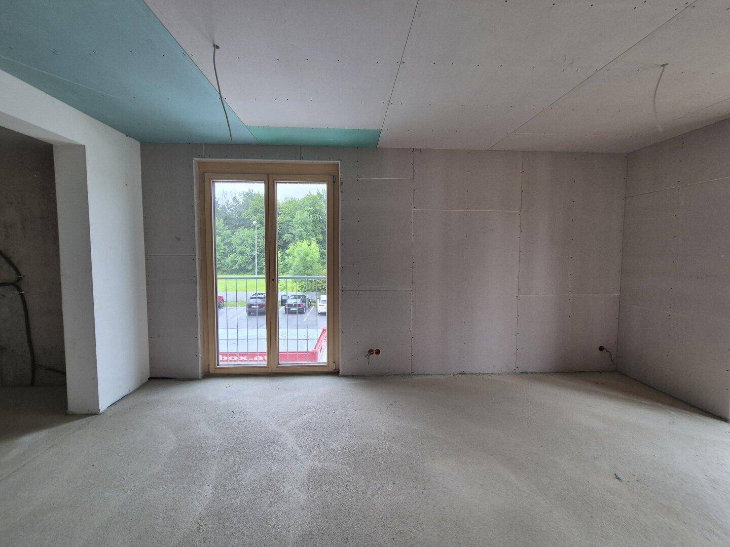 Rohbau eines Wohnbereichs mit Betonboden und Balkontür, die den Blick ins Grüne bietet.