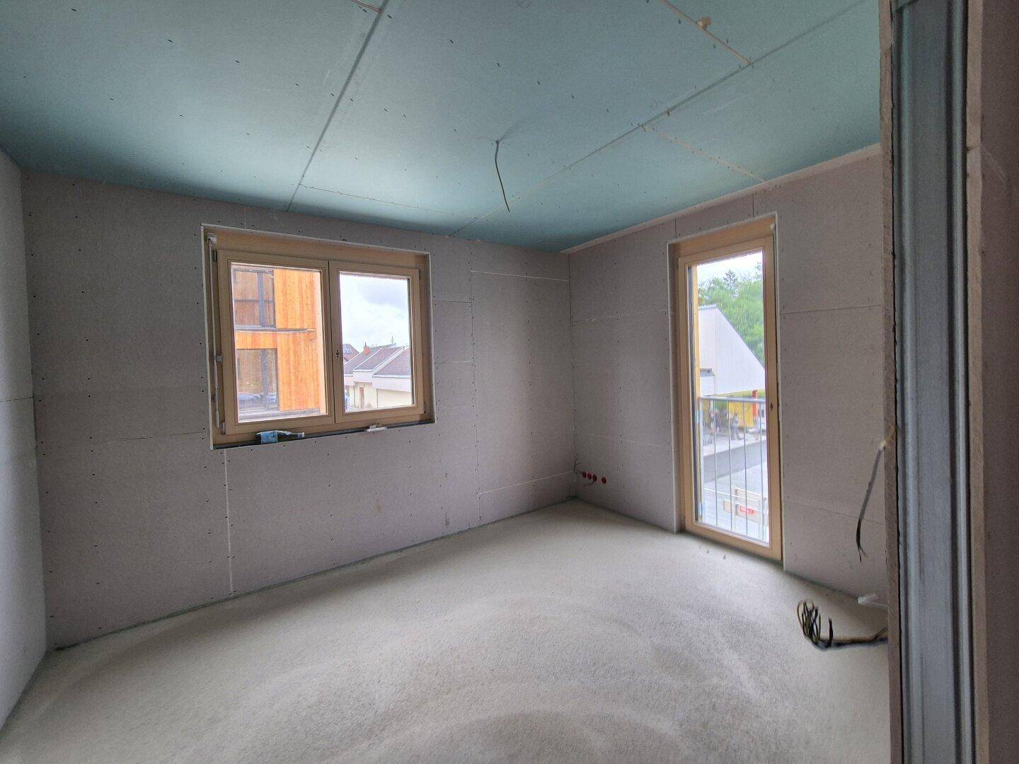 Rohbau eines Zimmers mit Betonboden, Fenster und Balkontür, bereit für den Ausbau.