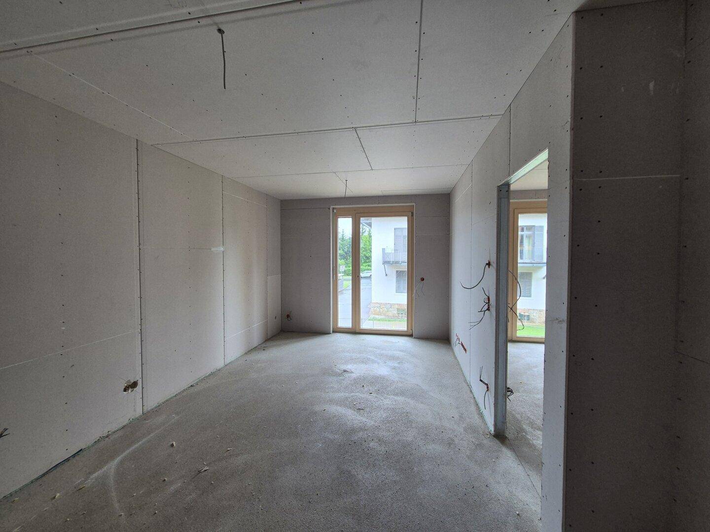 Leerer Raum im Rohbau mit Trockenbauwänden, Betonboden und Zugang zu einem Außenbereich.