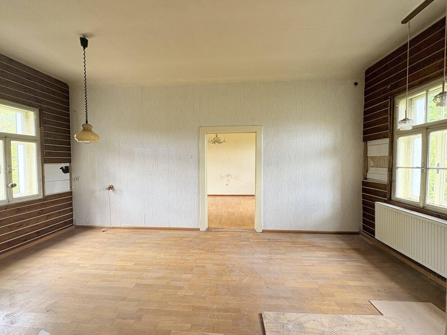 Geräumiger Raum mit hellem Parkettboden und teilweise holzvertäfelten Wänden, mit Durchgang zu einem weiteren Zimmer.