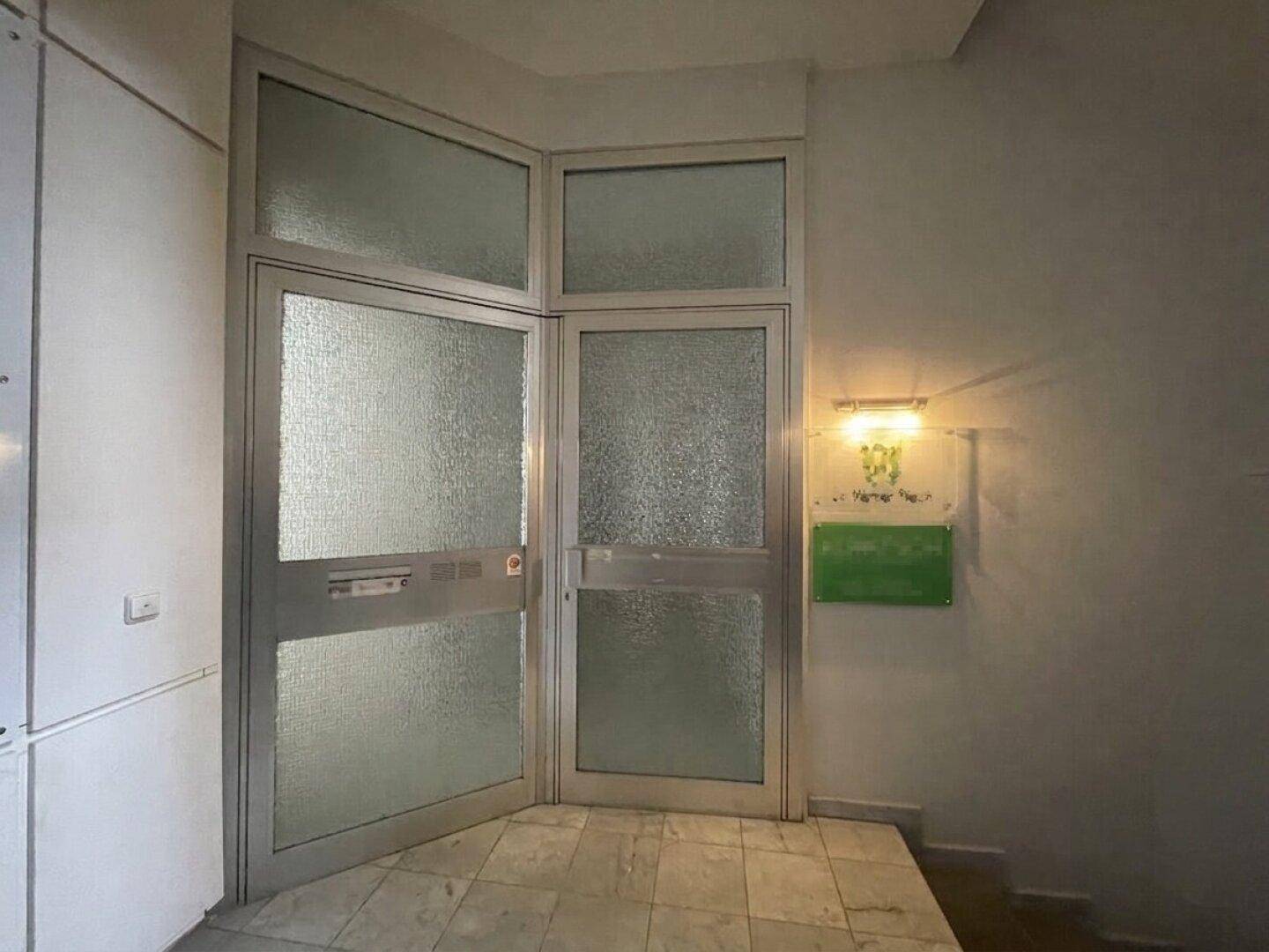Eingangsbereich mit verglaster Doppeltür, Fliesenboden und einer Wandleuchte, die den Raum erhellt.