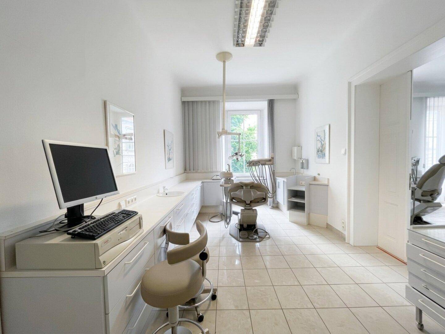 Behandlungszimmer mit Fliesenboden, medizinischem Stuhl und Arbeitsplatte mit Computerarbeitsplatz.