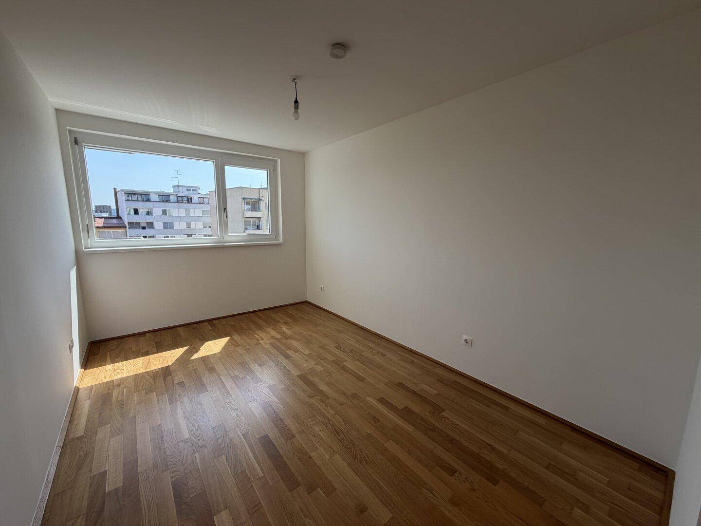 Leeres Schlafzimmer mit Holzboden und großem Fenster, das viel Tageslicht hereinlässt.