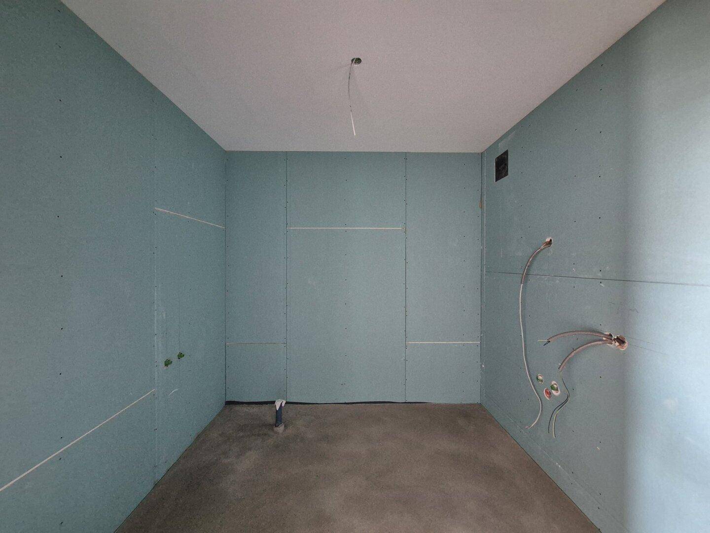 Badezimmer im Rohbauzustand mit feuchtigkeitsresistenten Wänden und sichtbaren Sanitäranschlüssen.
