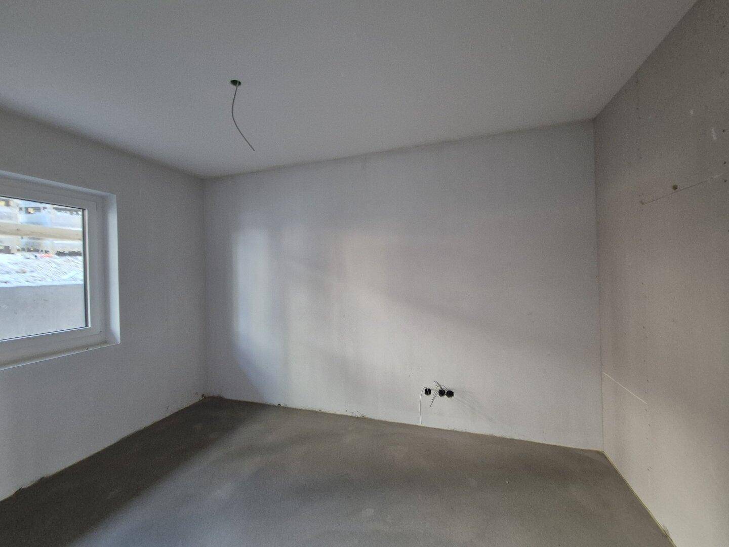 Rohbauzimmer mit Trockenbauwänden, Betonboden und einem Fenster, das natürliches Licht spendet.