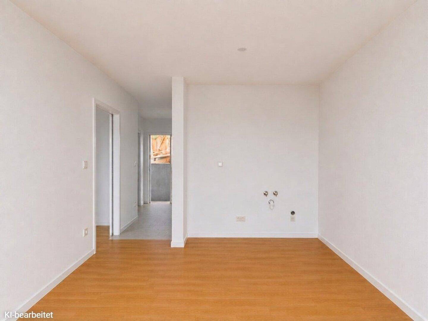 Heller Raum mit Holzboden und Anschlüssen für eine Küche, angrenzend an einen Flur.