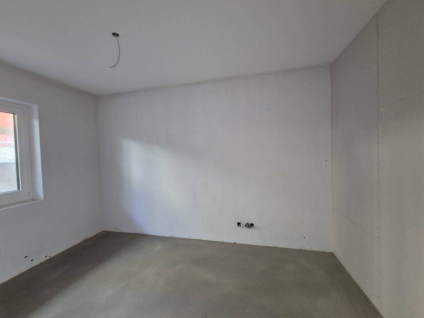 Unfertiger Raum mit Betonboden und einem Fenster, das natürliches Licht spendet.