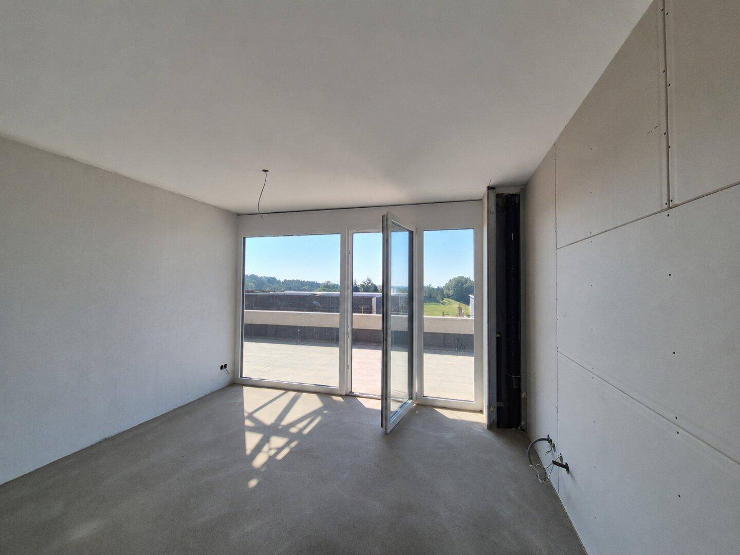 Unfertiger Raum mit Betonboden und großen Fenstern, die auf eine Terrasse führen.