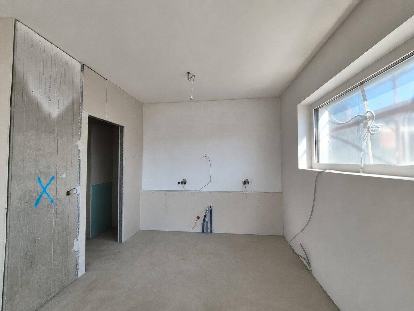 Unfertige Küche mit Rohbauwänden, Fenster und Anschlüssen für Spüle und Geräte.