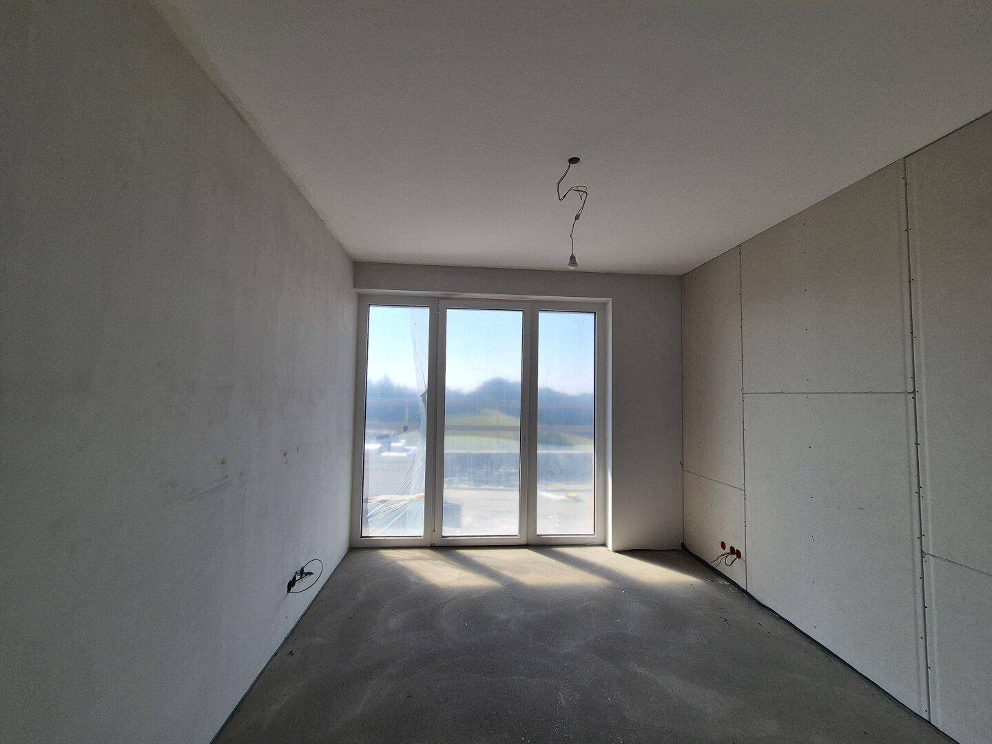 Unfertiger Raum mit großen Fenstern und Rohbauwänden, bereit für den weiteren Ausbau.