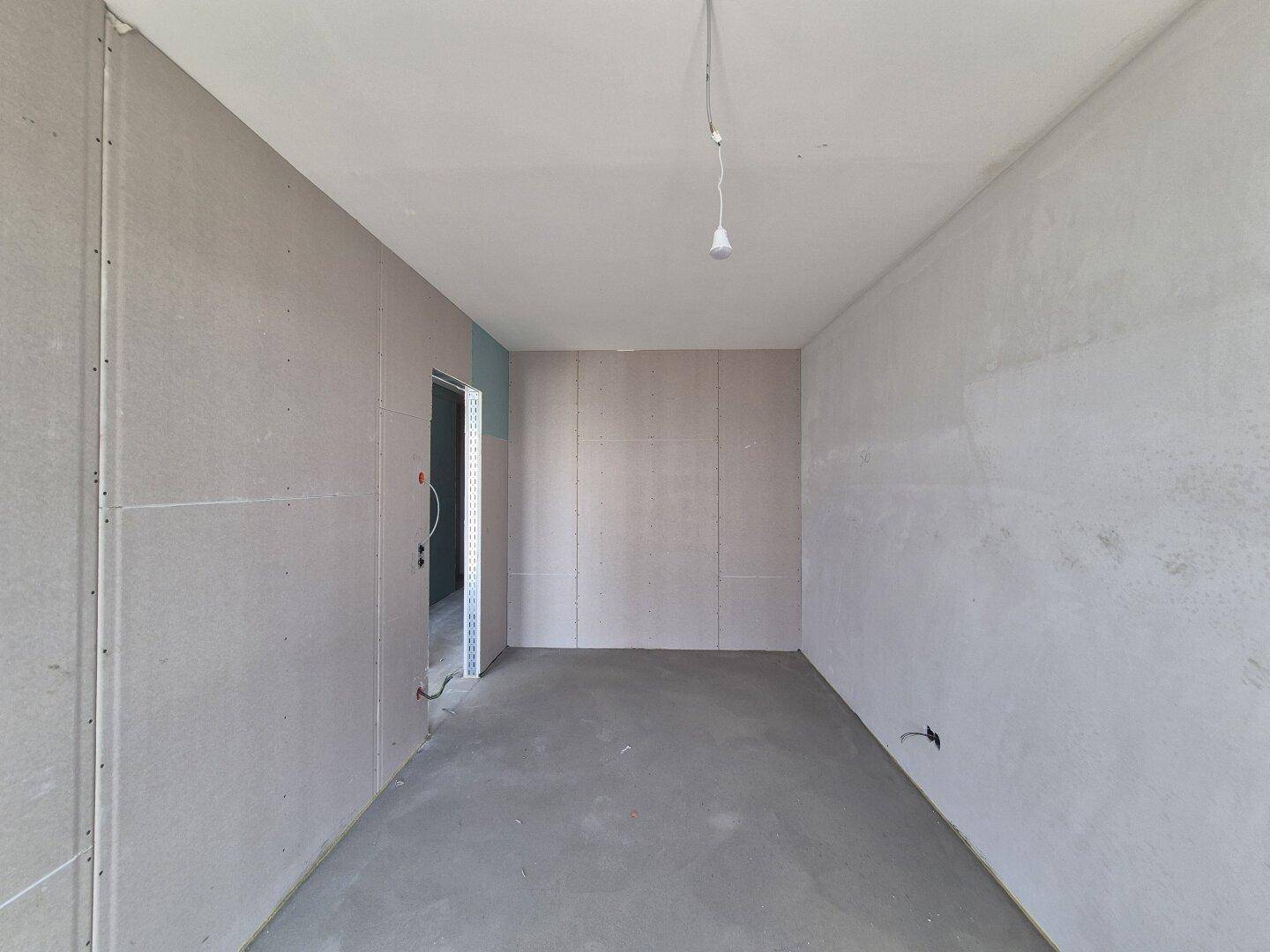 Unfertiger Raum mit grauen Trockenbauwänden und Betonboden, eine Glühbirne hängt von der Decke.