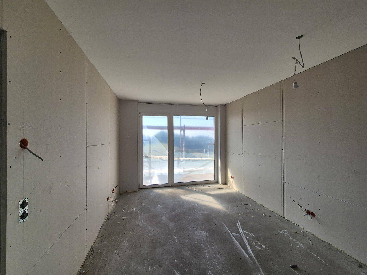 Unfertiger Raum mit Trockenbauwänden und Betonboden, der viel natürliches Licht durch ein großes Fenster erhält.