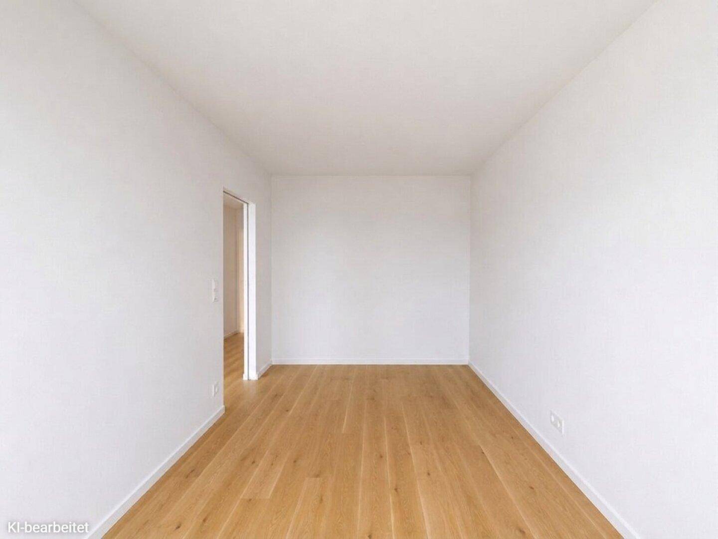 Leeres Zimmer mit hellen Holzböden und weißen Wänden, ein offener Durchgang führt in einen weiteren Raum.