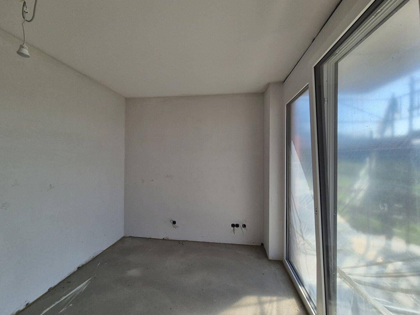 Rohbauzimmer mit Betonboden und großen Fenstern, bereit für den Innenausbau und die Gestaltung.