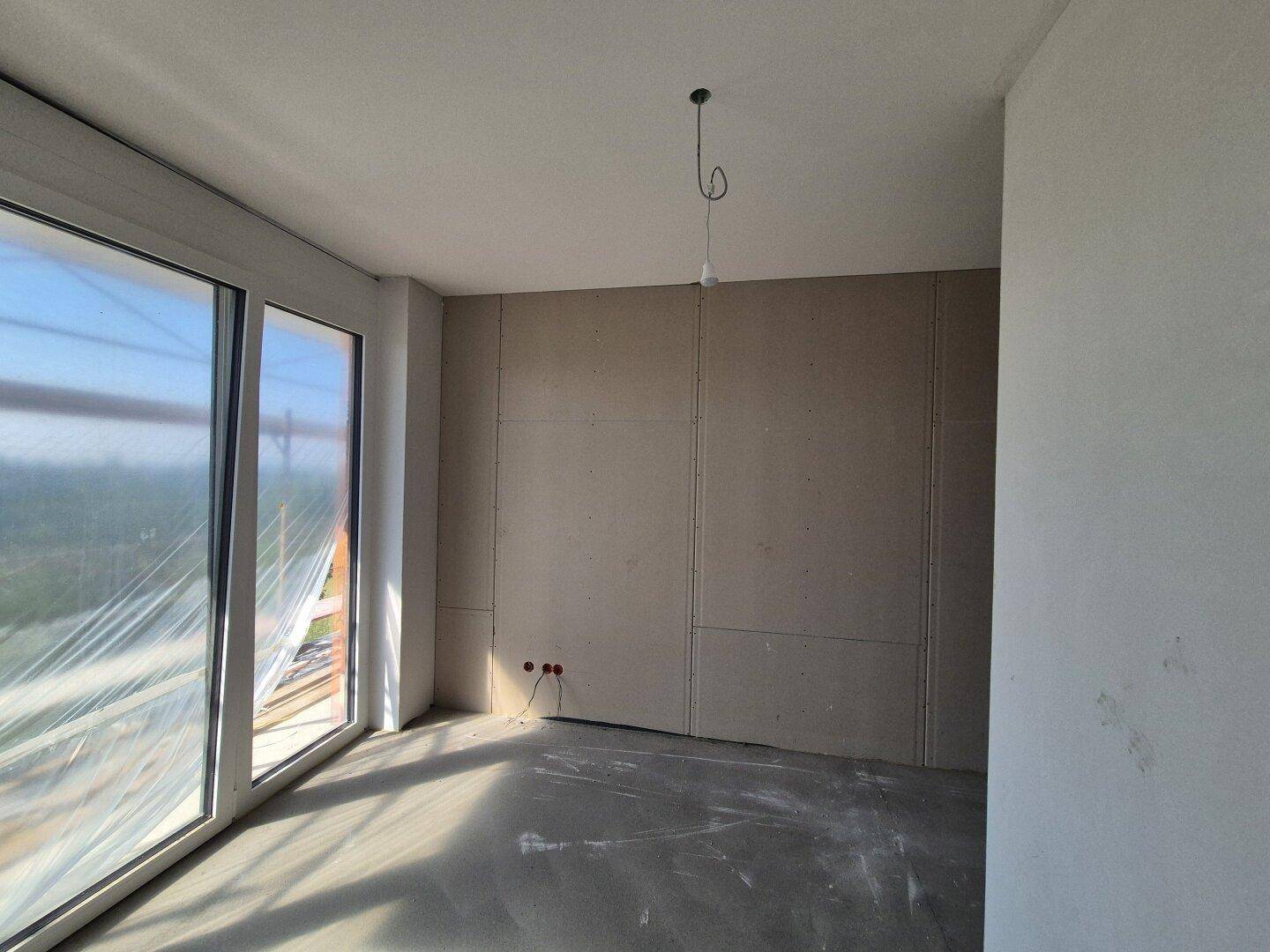Unfertiger Raum mit Trockenbauwänden und Betonboden, große Fenster bieten viel Tageslicht.