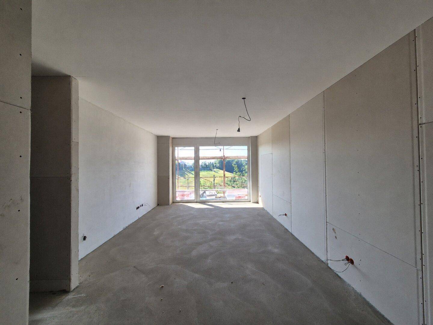 Geräumiger Rohbau mit Betonboden und Trockenbauwänden, große Fenster mit Blick ins Grüne.