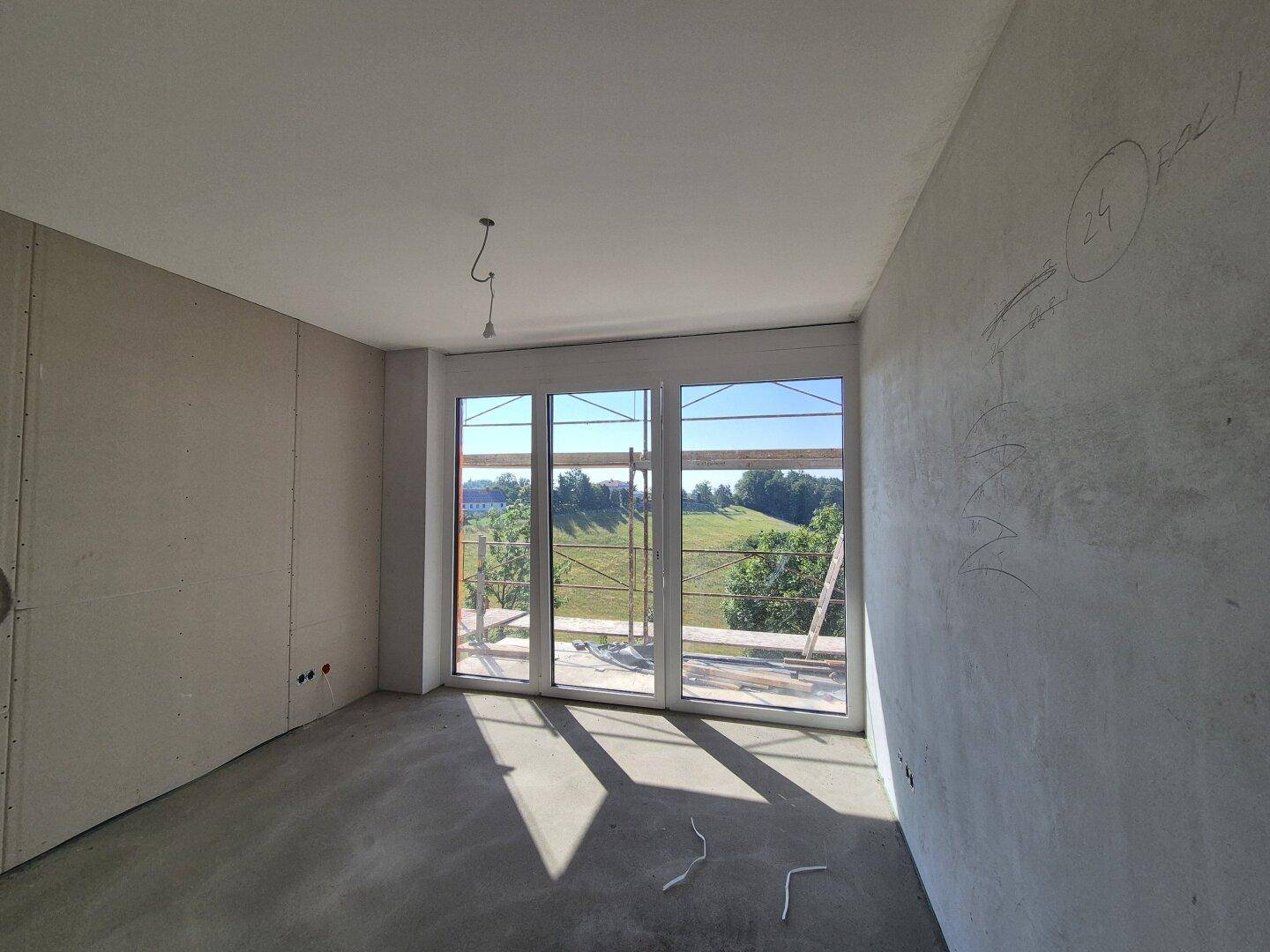 Raum im Rohbau mit Betonboden und großen Fenstern, die einen Blick ins Grüne bieten.