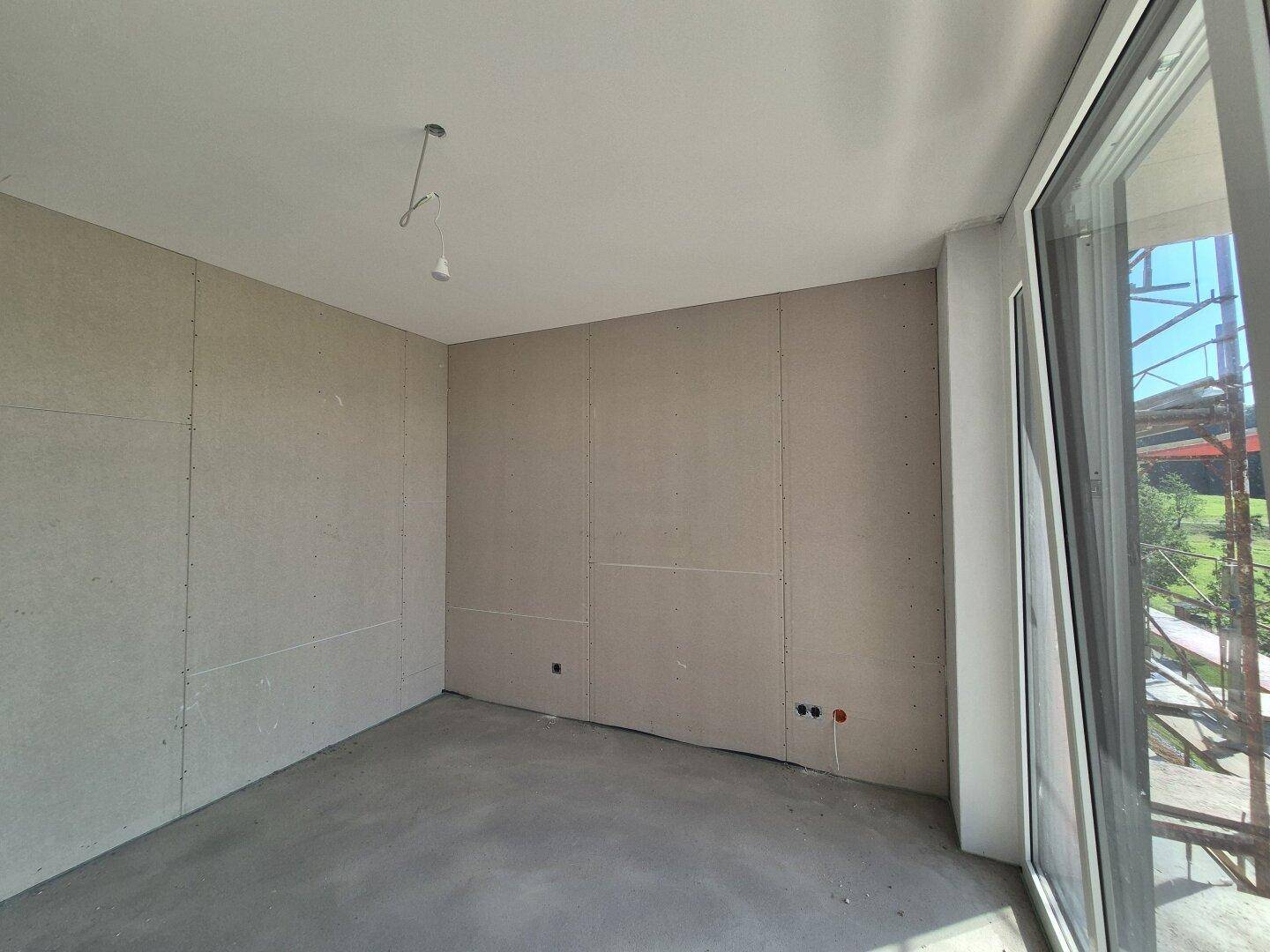 Raum im Rohbau mit Betonboden und großen Fenstern, die auf eine Baustelle blicken.