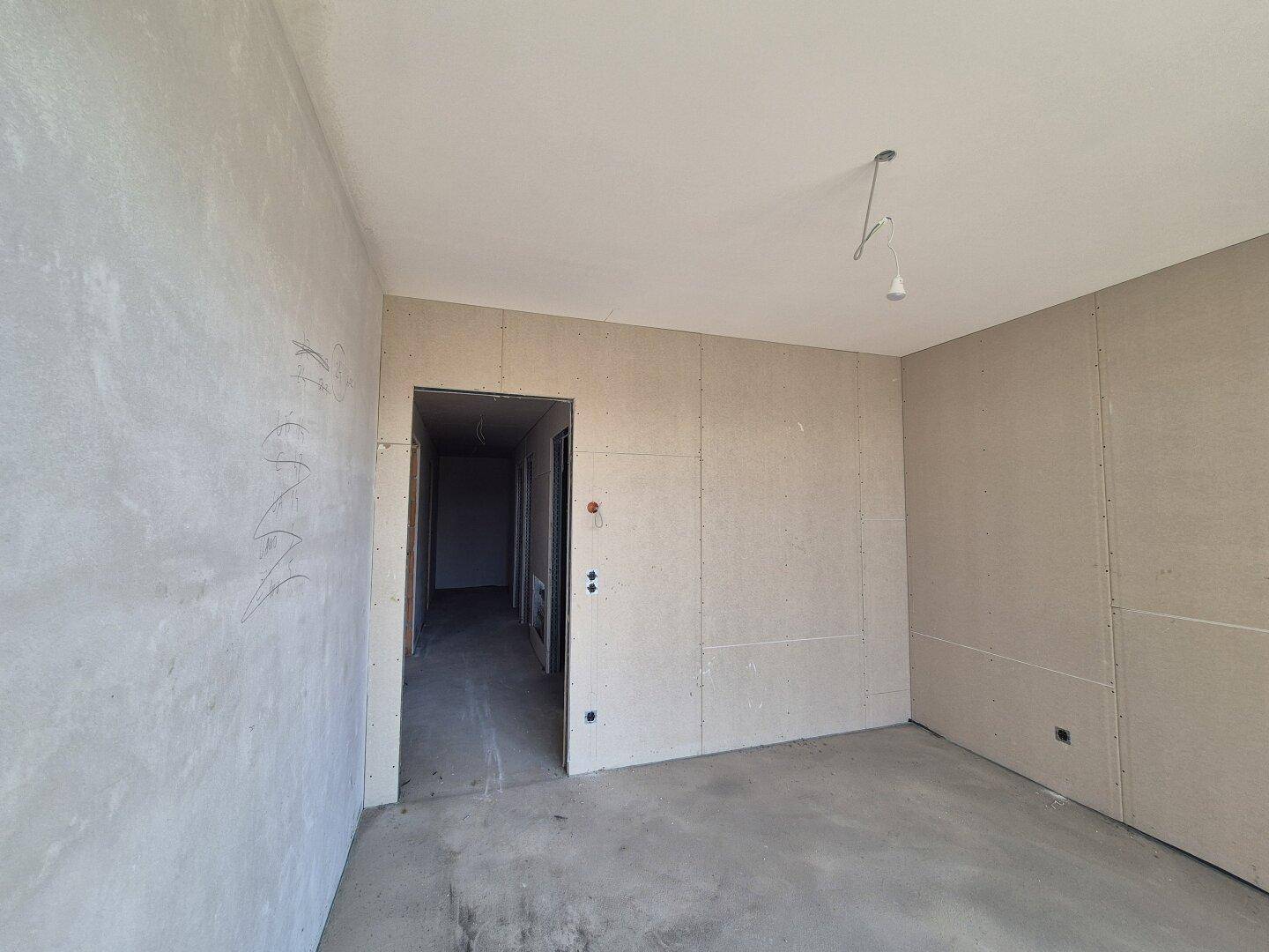 Rohbauzimmer mit Betonboden und Trockenbauwänden, Blick in einen Flur.