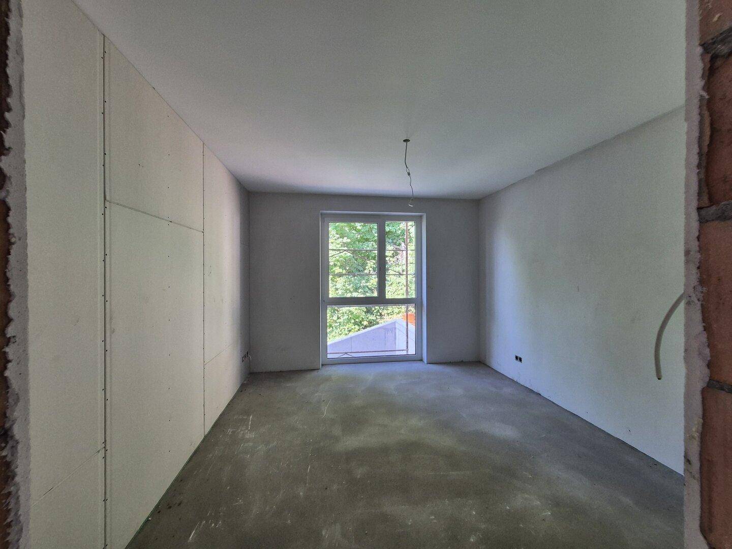 Rohbauzimmer mit Betonboden und Fenster, das einen Blick auf Bäume und Natur bietet.