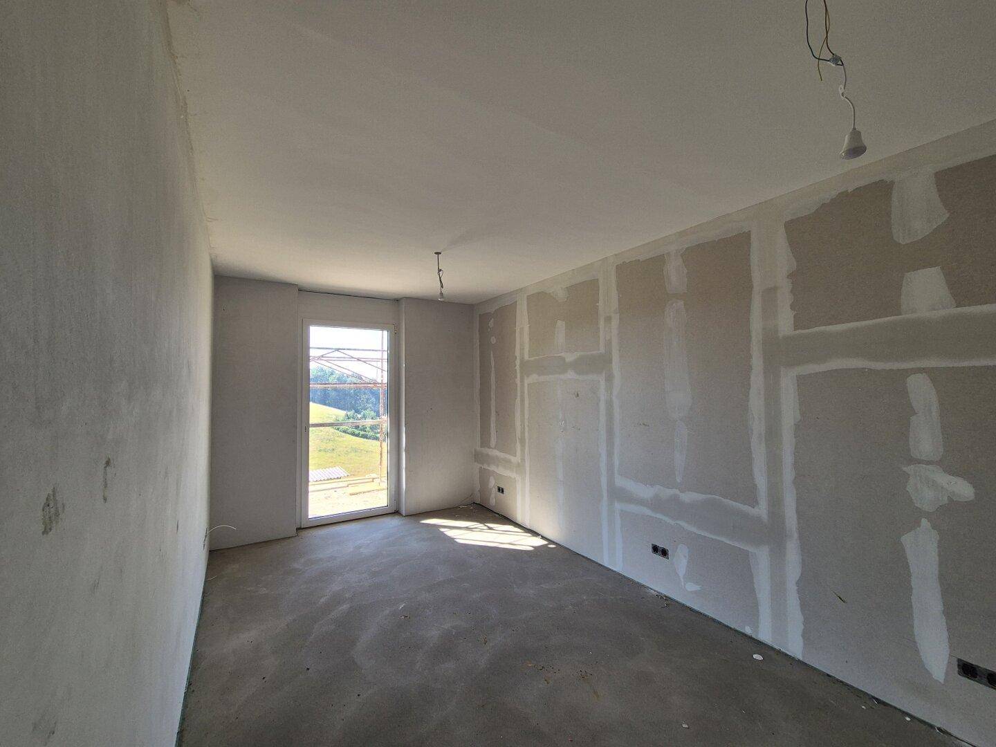 Raum im Rohbau mit Trockenbauwänden und Betonboden, mit einem Fenster für natürliches Licht.