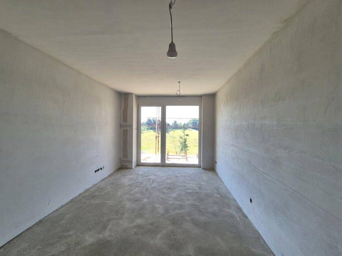 Unfertiger Raum mit großen Fenstern und Rohbetonboden, der einen weiten Blick ins Grüne bietet.