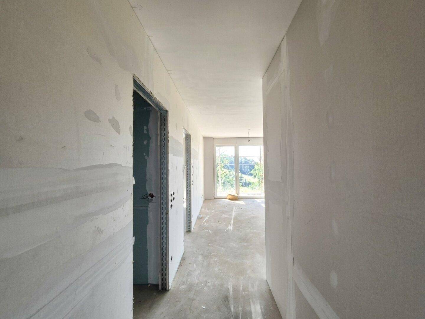 Unfertiger Flur mit unverputzten Wänden und Betonboden, der zu einem hellen Bereich führt.