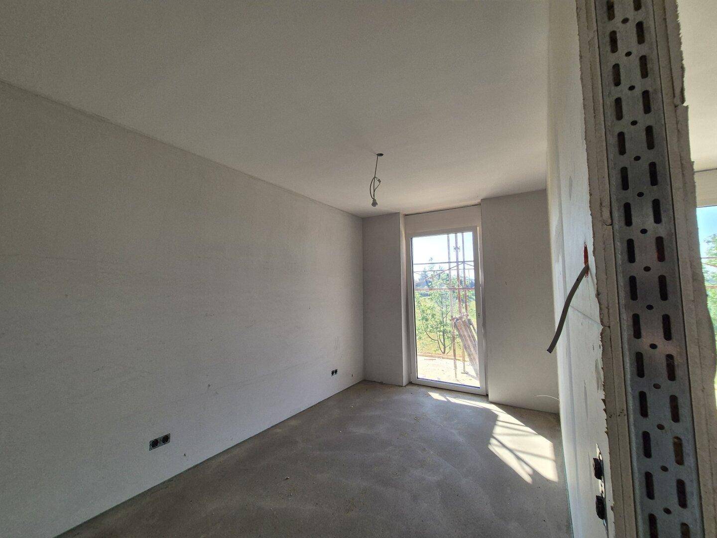 Unfertiges Zimmer mit Betonboden und einem Fenster, das einen Blick ins Grüne bietet.