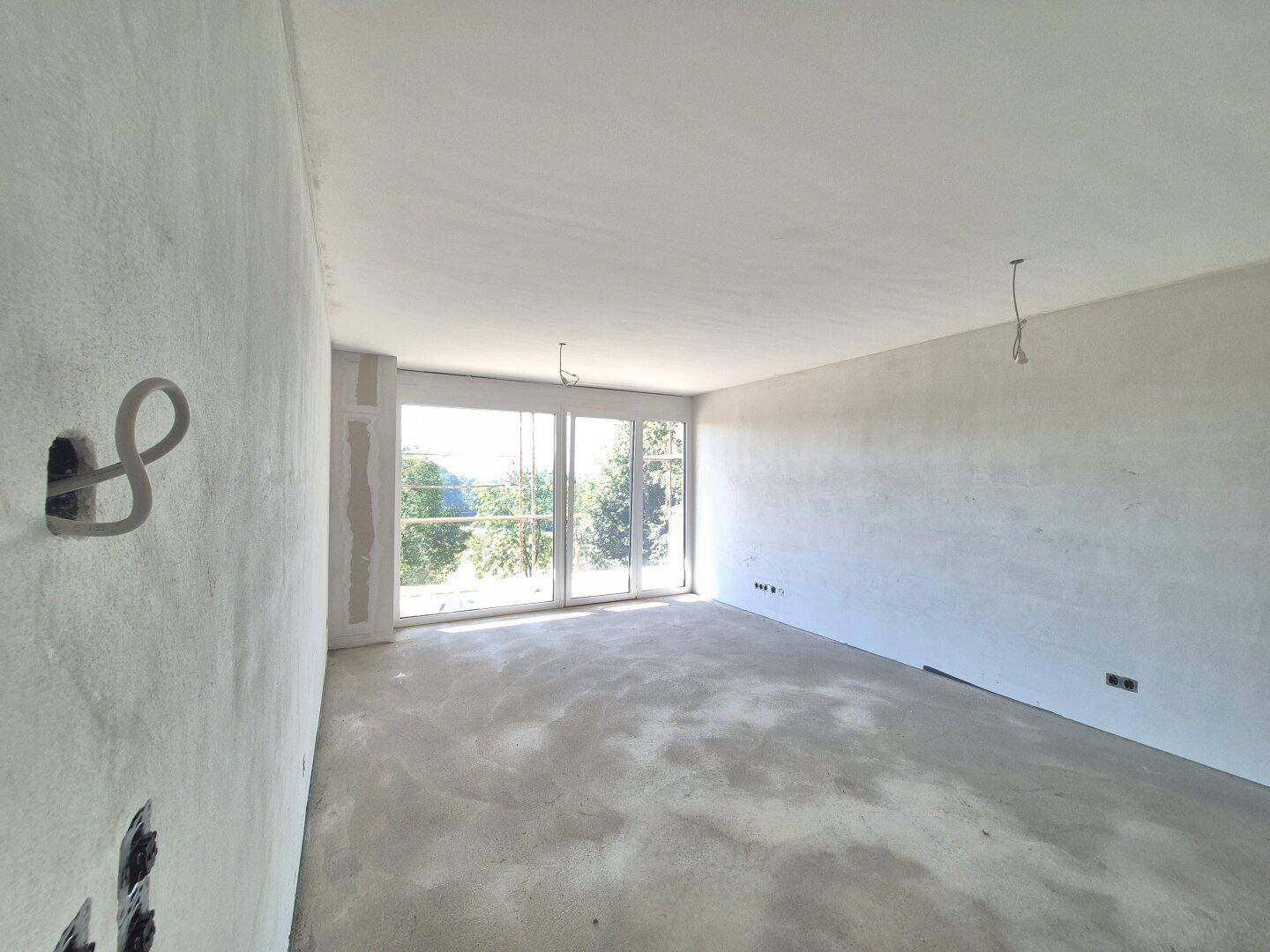 Rohbauzimmer mit Betonboden und großen Fenstern, die einen Blick auf die umliegende Natur ermöglichen.