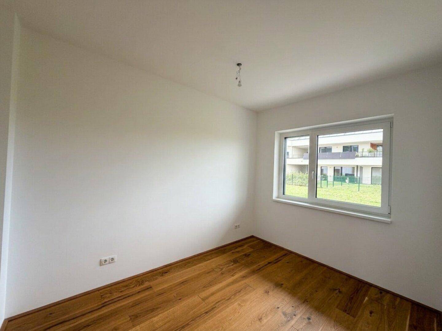 Helles, leeres Zimmer mit Holzboden und einem Fenster mit Blick auf die Nachbarschaft.